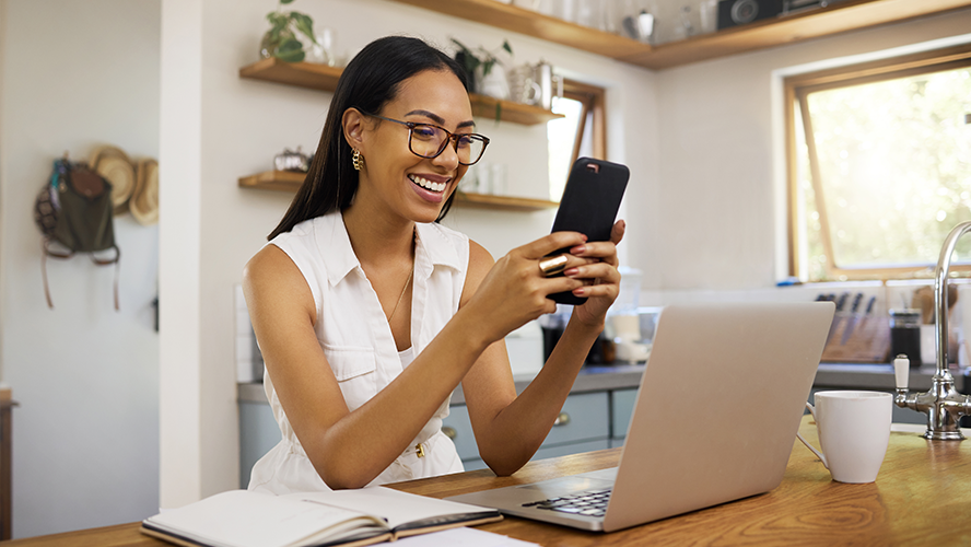 Woman at table with laptop looking at phone and smiling.